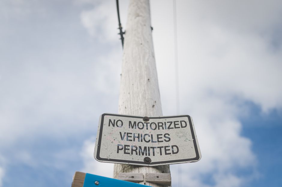 A weathered rectangular metal sign attached to a wooden utility pole with visible screws, displaying the message 'NO MOTORIZED VEHICLES PERMITTED' in black uppercase letters. The sign is positioned near the top of the pole, with a cloudy sky as the background. The utility pole has some cracks and minor wear, and there is a small, partially visible blue sign below the main sign, possibly indicating local regulations. The scene suggests a restricted access area, commonly encountered during house removal or relocation processes managed by companies like Man With a Van Bickley, as part of the planning around house removals and furniture transport in compliance with local permit rules.