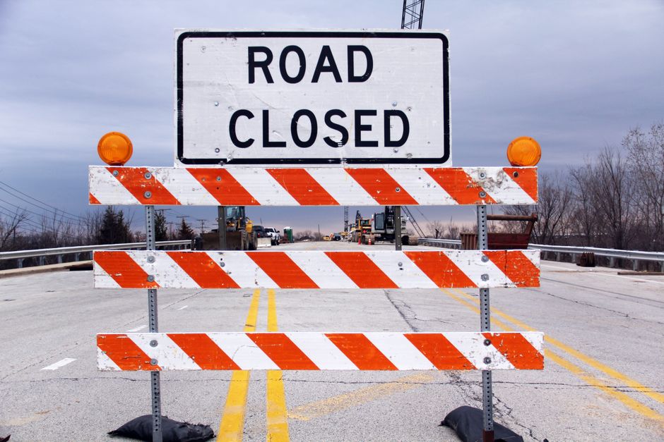 A road closed barrier with red and white reflective stripes, supported by metal stands anchored with sandbags on either side, obstructs a two-lane street during daytime with overcast sky. Behind the barrier, there is a large white sign reading 'ROAD CLOSED' in bold black letters. The street surface is asphalt with yellow double lines in the center, and there is minimal traffic visible further down the road, including construction vehicles and equipment. On the left side, there is a metal guardrail, and in the background, leafless trees and utility poles are visible, indicating a winter season. The scene appears to be part of a road closure due to construction or maintenance, which could impact house removals or relocation services operating in the area. Man With a Van Bickley occasionally works in similar environments, assisting clients with furniture transport and home relocation logistics, particularly when access is restricted by road closures or permit rules related to local traffic regulations.