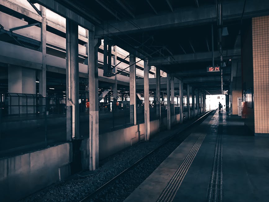 A train station platform with a partially covered area featuring concrete and metal structural supports. The platform appears to be quiet with minimal passengers visible, some small figures are seen in the distance near the edge of the platform, while a person is walking away from the camera towards the illuminated exit. Alongside the platform, there are tactile paving strips for accessibility and a digital clock displaying the time as 15:14. The station environment is dimly lit, with natural light coming from the open exit area at the end of the platform. This scene exemplifies a typical urban railway station, suitable for house removals or property relocation services seeking transportation or logistical support through the rail network, as offered by Man With a Van Bickley.