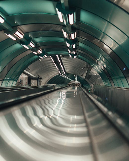 A photograph taken from the low perspective of an escalator inside a modern underground station shows the metal surface and moving steps leading into a tunnel-like structure. The ceiling features curved, metallic panels in a greenish hue with integrated LED lighting strips that provide illumination along the length of the tunnel. The escalator's handrails are visible along the sides, and the polished metal surface reflects the overhead lights, creating a pattern of light and shadow. The setting appears clean and well-maintained, with an industrial design typical of urban public transport infrastructure. This image captures the smooth, continuous flow of movement within an underground station environment, relevant to urban transportation and the logistics involved in relocating or moving between city locations, such as those handled by companies like Man With a Van Bickley.