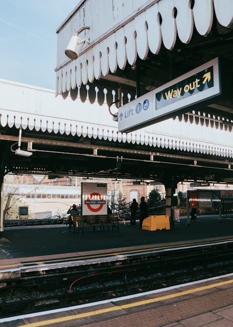 A train station platform with a partially covered area featuring concrete and metal structural supports. The platform appears to be quiet with minimal passengers visible, some small figures are seen in the distance near the edge of the platform, while a person is walking away from the camera towards the illuminated exit. Alongside the platform, there are tactile paving strips for accessibility and a digital clock displaying the time as 15:14. The station environment is dimly lit, with natural light coming from the open exit area at the end of the platform. This scene exemplifies a typical urban railway station, suitable for house removals or property relocation services seeking transportation or logistical support through the rail network, as offered by Man With a Van Bickley.
