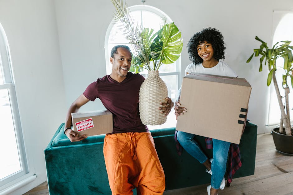 A man and a woman, both smiling, hold packing boxes inside a bright living room with natural light coming through arched windows. The man, dressed in a burgundy t-shirt and orange pants, carries a small cardboard box labeled 'Fragile' in red, while the woman, wearing a white t-shirt and jeans, holds a larger cardboard box. Between them, the man also holds a large ceramic vase with green tropical leaves, showcasing part of the packing and unpacking process involved in house removals. Behind them, a dark green sofa and a tall potted plant are visible, with the room featuring light-colored walls and wooden flooring. The scene captures the preparatory phase of a home relocation, highlighting furniture and box transportation, as part of the comprehensive moving services provided by Man With a Van Bickley. This image exemplifies packing, loading, and transport activities typical of professional removals, with natural indoor lighting enhancing the clarity of the objects and environment.