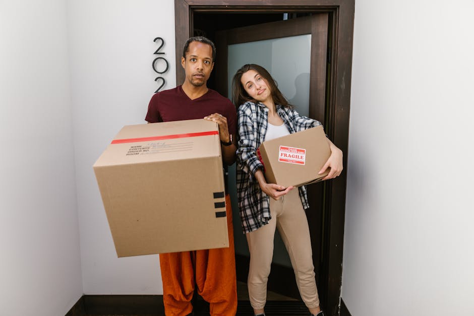 A man and a woman, both smiling, hold packing boxes inside a bright living room with natural light coming through arched windows. The man, dressed in a burgundy t-shirt and orange pants, carries a small cardboard box labeled 'Fragile' in red, while the woman, wearing a white t-shirt and jeans, holds a larger cardboard box. Between them, the man also holds a large ceramic vase with green tropical leaves, showcasing part of the packing and unpacking process involved in house removals. Behind them, a dark green sofa and a tall potted plant are visible, with the room featuring light-colored walls and wooden flooring. The scene captures the preparatory phase of a home relocation, highlighting furniture and box transportation, as part of the comprehensive moving services provided by Man With a Van Bickley. This image exemplifies packing, loading, and transport activities typical of professional removals, with natural indoor lighting enhancing the clarity of the objects and environment.