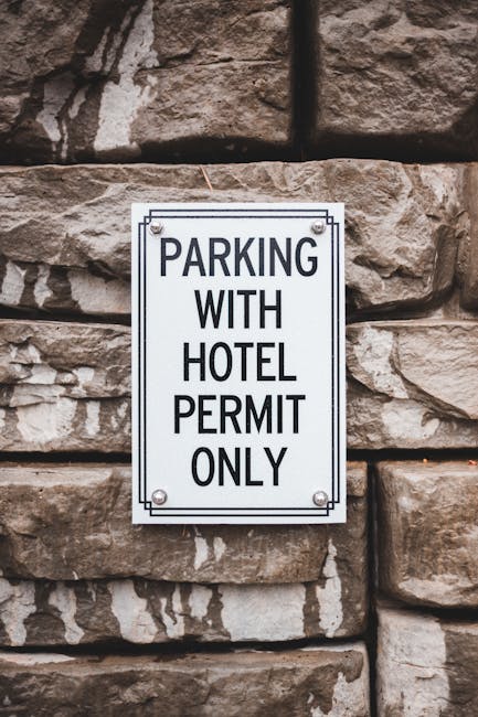 Close-up view of a rectangular white metal sign with black text stating 'PARKING WITH HOTEL PERMIT ONLY', mounted on a textured brown stone wall. The sign is secured with four screws, one at each corner. In the background, part of a street or driveway area can be seen, suggesting the location is adjacent to a building entrance or parking area. This image relates to house removals and moving services, highlighting parking permit considerations for home relocation or furniture transport—an aspect managed by companies like Man With a Van Bickley when planning local moves and ensuring proper parking arrangements.