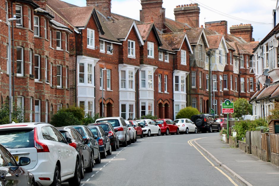 A residential street lined with traditional red-brick terraced houses featuring pitched roofs, white-framed sash windows, and some with small front gardens. Parked along the curb are various cars, including hatchbacks, sedans, and SUVs, all facing in the same direction. The street curves gently to the right, with double yellow lines marking the parking restrictions. On the pavement, there are a few green bushes and a street lamp visible. A sign indicating parking regulations is mounted on a post next to the sidewalk. The environment is well-lit with natural daylight, and there are no pedestrians or moving vehicles in the scene. This setting illustrates a typical urban area suitable for home relocation and furniture transport, often requiring careful parking management and moving logistics. A moving company like Man With a Van Bickley would coordinate loading and unloading in such residential streets as part of their house removals services.