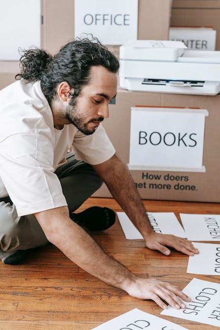 A man with curly dark hair and a beard, dressed in a white shirt and dark trousers, is kneeling on a wooden floor inside a room during a house removal process. He is carefully arranging large sheets o