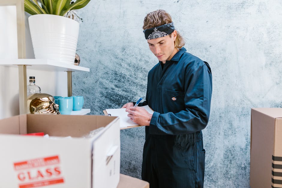 A man with curly dark hair and a beard, dressed in a white shirt and dark trousers, is kneeling on a wooden floor inside a room during a house removal process. He is carefully arranging large sheets o
