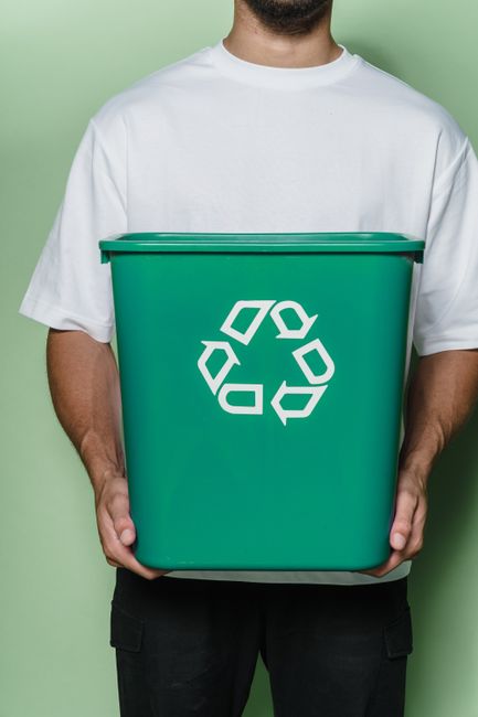 A person wearing a plain white T-shirt holding a large green recycling bin with a white recycling symbol on the front, standing against a solid light green background. The bin is made of plastic, with a flat lid, and the person grips it with both hands, one on each side. The image captures the upper body and hands of the individual, emphasizing environmentally responsible waste disposal, which aligns with sustainable home relocation practices. The scene is well-lit, with no visible clutter or background distractions, highlighting the focus on recycling in the context of house removals and responsible disposal services offered by Man With a Van Bickley.