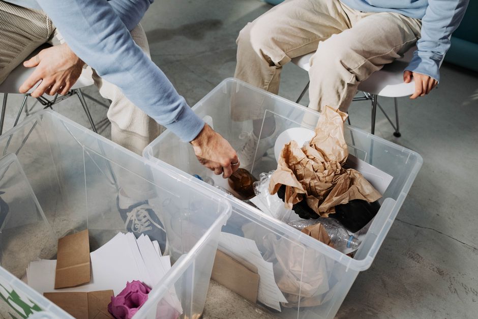 Two individuals are engaged in packing or sorting items inside a home during a house relocation process. One person, seated on the left, is wearing light grey pants and a blue long-sleeve shirt, and is reaching into a clear plastic storage bin filled with crumpled brown paper, plastic bottles, and other packing materials. The second person, seated on the right, is dressed in beige trousers and a light blue shirt, with their hands resting on their knees as they observe. The containers are situated on a concrete floor, and the environment appears to be an indoor space such as a garage or storage area. Visible objects in the bins include folded white and brown paper packages, plastic bottles, and packing paper, indicating the process of packing household belongings for a move. This activity is indicative of the packing and moving preparations undertaken by professionals like Man With a Van Bickley, who specialise in removals and home relocations, ensuring safe handling and transportation of possessions.