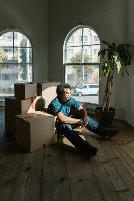 A man with curly hair, wearing a blue shirt and dark trousers, is sitting on the wooden floor of a room with high ceilings and large, arched windows, which allow natural daylight to illuminate the space. Behind him are several cardboard boxes secured with red tape, indicating they are prepared for moving. The boxes are stacked and positioned near a large, leafy potted plant placed against the white wall. The room appears to be part of a residential property, possibly undergoing furniture transport or packing during a home relocation. The windows reveal a street view with parked cars and a building across the road. The scene depicts a moment during the loading process, where the individual is resting amidst packing materials, consistent with the context of a professional removals service such as those offered by Man With a Van Bickley, supporting emergency eviction moves or same-day house removals.