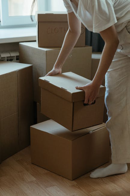 A person wearing light-colored clothing is seen lifting and stacking cardboard boxes inside a room, preparing for home relocation. The boxes vary in size, with some labeled 'CLOTHES,' and are placed on a wooden floor near a window that provides natural lighting. Additional cardboard boxes are visible in the background, some on the floor and others positioned on furniture or flat surfaces. The individual is in the process of packing or unpacking as part of a furniture transport or moving process. This scene illustrates the careful handling of packing materials typical in professional removals, such as those offered by Man With a Van Bickley, supporting efficient and organized unloading or loading during a house move.