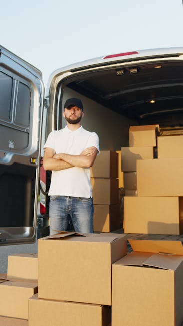 A man wearing a black cap, white t-shirt, and jeans stands with arms crossed outside the open rear doors of a van, which is filled with neatly packed cardboard boxes of various sizes stacked on the vehicle's floor and sides. The boxes are made of plain brown corrugated cardboard, some with flaps open revealing additional packing materials inside. The van is parked on a paved area next to a building, suggesting a home relocation or moving process. Nearby, additional boxes are placed on the ground, ready for loading or unloading. The scene is under natural daylight, indicating bright weather, and the interior of the van is illuminated by built-in lights. This image, associated with Man With a Van Bickley, depicts the logistics of furniture transport and packing during a professional removals service, supporting efficient and organized home moving activities.