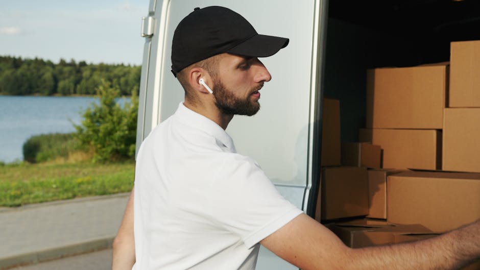A male removal worker wearing a black cap, white t-shirt, and wireless earbuds is seen loading cardboard boxes into the back of a large white van. The boxes are stacked neatly inside the vehicle, which is positioned on a paved surface near a body of water with green trees in the background. The worker's right arm is extended to place or adjust a box on the stack. The scene is outdoors during daylight, with natural lighting highlighting the loading process typical of home relocation and furniture transport operations conducted by Man With a Van Bickley. The image captures the careful handling of packing materials as part of professional removals services, emphasizing efficient logistics in local house moves.
