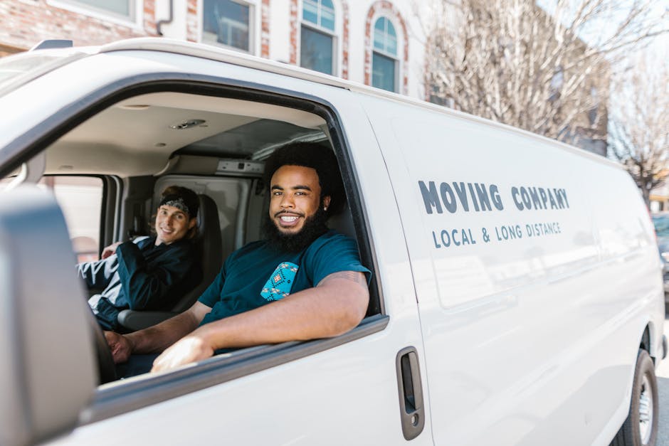 Inside a white moving van parked on a residential street near Bickley Village, two movers from Man With a Van Bickley are visible. The driver, a young man with dark curly hair and a beard, is seated in the cab with the window rolled down, smiling at the camera. Next to him, a young woman with dark hair, wearing a black jacket and a headband, is seated inside the van, also smiling. The interior of the van shows various packing materials such as cardboard boxes, some wrapped in plastic film, and blankets, indicating an ongoing home relocation or furniture transport. The van's side panel displays the company’s branding, including the words “MOVING COMPANY” and “LOCAL & LONG DISTANCE” service. The background includes the exterior of residential buildings and leafless trees, suggesting a clear day suitable for loading or unloading household items. The scene captures the loading process part of the packing and moving service provided by Man With a Van Bickley, supporting efficient house removals.