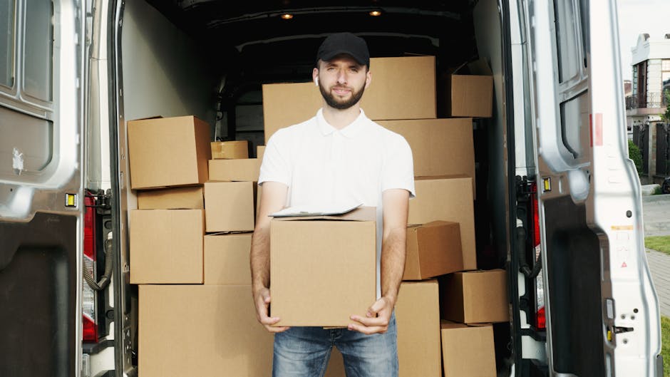A man with a beard, wearing a white polo shirt, jeans, and a black cap, stands inside the back of a large white van with its rear doors open. He is holding a medium-sized cardboard box, which is part of a collection of similarly packed cardboard boxes stacked neatly inside the van. The boxes vary in size and are sealed with packing tape. The van is parked outside a residential area on a paved street, with part of a house and greenery visible in the background. The interior of the van is empty apart from the packed boxes, and the scene suggests a home relocation or moving process. Man With a Van Bickley specializes in furniture transport and packing and loading services, supporting efficient house removals near Bickley Village.