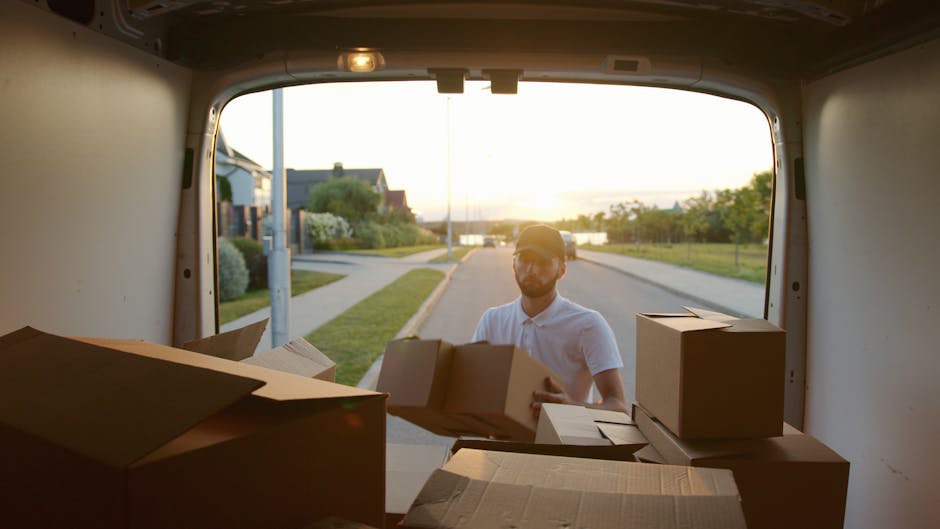 Inside a white moving van parked on a residential street near Bickley Village, two movers from Man With a Van Bickley are visible. The driver, a young man with dark curly hair and a beard, is seated in the cab with the window rolled down, smiling at the camera. Next to him, a young woman with dark hair, wearing a black jacket and a headband, is seated inside the van, also smiling. The interior of the van shows various packing materials such as cardboard boxes, some wrapped in plastic film, and blankets, indicating an ongoing home relocation or furniture transport. The van's side panel displays the company’s branding, including the words “MOVING COMPANY” and “LOCAL & LONG DISTANCE” service. The background includes the exterior of residential buildings and leafless trees, suggesting a clear day suitable for loading or unloading household items. The scene captures the loading process part of the packing and moving service provided by Man With a Van Bickley, supporting efficient house removals.