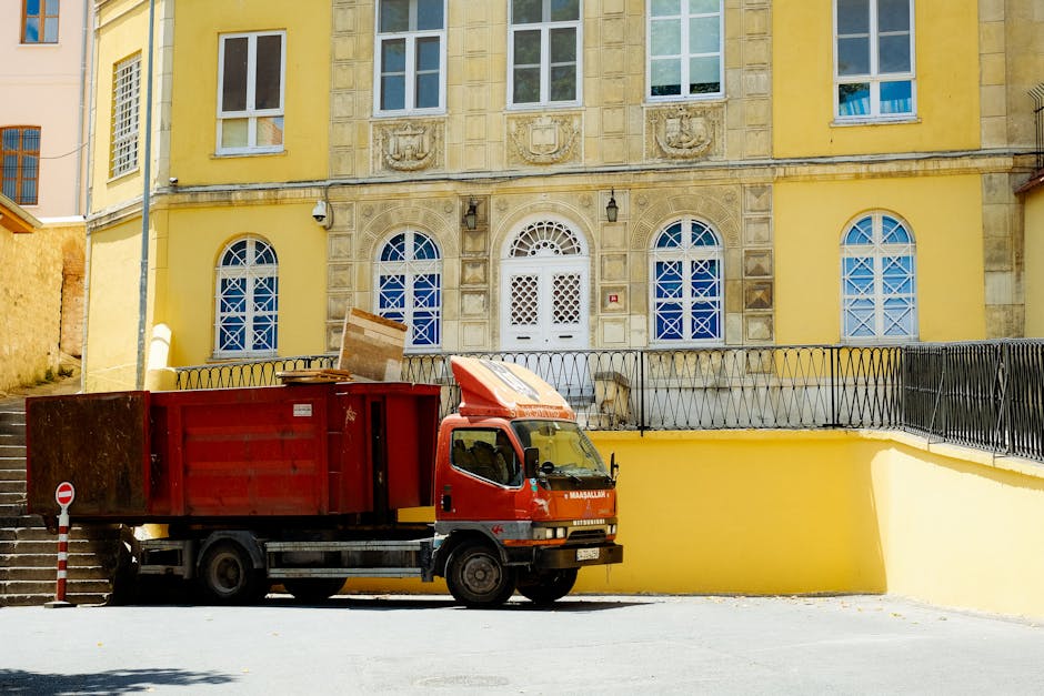A man wearing a black cap, white polo shirt, and jeans is sitting on a paved surface, surrounded by several cardboard boxes used for moving. He is carefully writing or signing on a piece of paper plac
